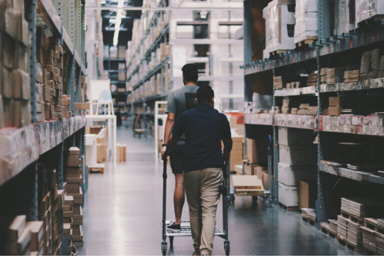 Two men pushing a cart through a warehouse, filled with shelves and boxes.